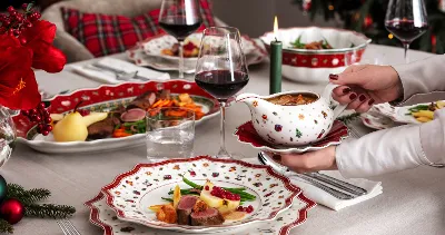 A festive dining table setting with Christmas dinnerware from Villeroy & Boch Toy’s Delight and presents plates of meat, vegetables, and sauce. A person holds a gravy boat, glasses of red wine, and a lighted candle.