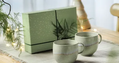 Two green Perlemor cups on a table with a matching green gift box in the background, decorated with a tree and lights.