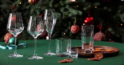 A stylish arrangement from the Toys Delight glassware collection by Villeroy & Boch, consisting of wine glasses and drinking glasses, elegantly placed on a green table. Dried citrus slices lie on a wooden tray in the foreground, complemented by the festive presence of a decorated Christmas tree in the background.