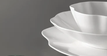 Three white Pura plates with scalloped edges are placed on a grey surface. Three white Pura plates with serrated edges are stacked on a grey surface next to a round, green object.
