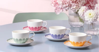 Vier Fleur Teetassen mit passenden Untertassen Vier Fleur Teetassen mit passenden Untertassen in unterschiedlichen Pastellfarben sind auf einem blauen Tisch neben Vasen mit rosa und weißen Blumen angeordnet.