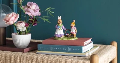 Two decorative bunny figurines from the Bunny Tales collection by Villeroy & Boch on stacked books on a wicker bench, next to a vase with ranunculus and roses.
