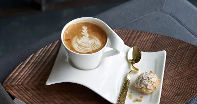 A Villeroy & Boch NewWave latte cup with latte art on a wavy white saucer is combined with a small cookie and a golden spoon, all beautifully arranged on a wooden tray.