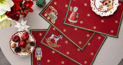Red placemats with Christmas motifs on a round table with festive plates, cookies, fruit, nuts, and ornaments.