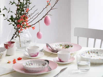 A bright, springtime dining table set with pastel-colored dinnerware from the Rose Garden and Spring Fantasy collections, decorated with floral patterns and Easter eggs hanging from a branch as a centerpiece.
