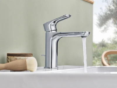 En moderne kromarmatur med rennende vann som er installert på en hvit O.novo-servant. Ved siden av står det en børste og en trebeholder.