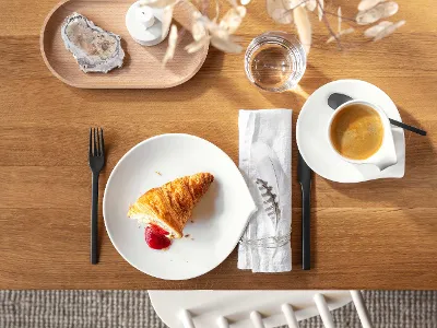 Top view of a wooden table with a croissant and jam on a white Flow plate, a cup of coffee, cutlery, a napkin, a glass of water and decorative elements.