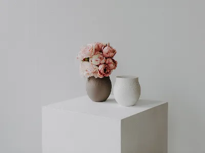 Un vase marron avec des fleurs roses et un vase blanc en céramique sont posés sur un cube blanc sur un fond clair et sobre.