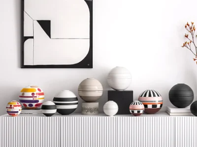 A series of patterned spherical tableware sets on a white shelf A series of patterned spherical tableware sets on a white shelf, underneath a geometric black-and-white painting featuring a vase with orange flowers.