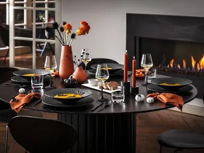 Une table de salle à manger moderne dressée pour cinq personnes avec de la vaisselle Manufacture noire, des serviettes orange et des décorations automnales, dont des vases à fleurs et des bougies allumées, à côté d’une cheminée où brûle un feu.