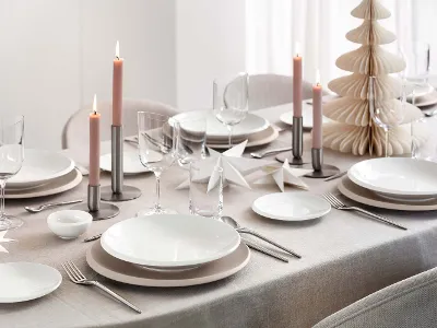 A beautifully set table with white NewMoon dinnerware, wine glasses, pink candles, silver flatware, and paper decorations on a beige tablecloth. There is also a small paper Christmas tree.