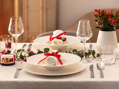 Elegant dining table for two with white Manufacture plates, napkins with red ribbons, wine glasses, silver flatware, and decorative plants.