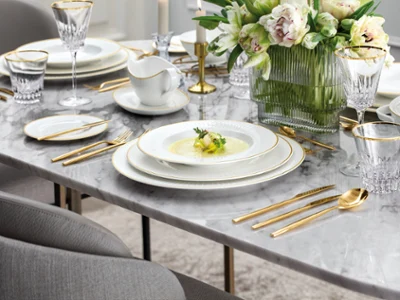 Elegant table set on a marble surface with gold-rimmed plates, gold flatware, crystal glasses, a vase of white flowers, and a soup served on the central plate.