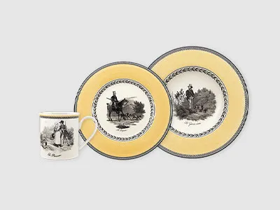 Eine Tasse und zwei Teller mit gelbem Rand und schwarzen Illustrationen pastoraler Szenen auf hellgrauem Hintergrund.