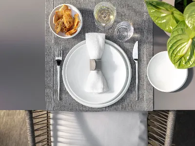 Draufsicht auf ein Gedeck mit einem weißen NewMoon Tellern, einer Serviette, Besteck, einer Müslischale, einem Glas und einer Pflanze auf einem gewebten Tischset.