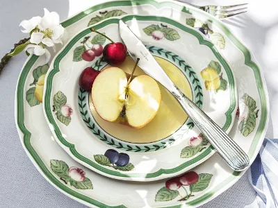 A table setting with a stack of French Garden plates from Villeroy & Boch with a floral pattern, a sliced apple, two cherries, a knife, and a fork. Next to it are white flowers and a blue ribbon.