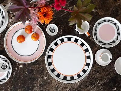 Memphis Tischgedeck von Villeroy & Boch mit dekorativen Tellern, einem Becher, Tassen und Pflaumen auf einer mit leuchtenden Blumen geschmückten braunen Marmoroberfläche.