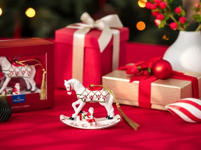 Festive Christmas scene with a white rocking horse ornament, red and gold gifts, decorative baubles, and a vase with holly on a red tablecloth