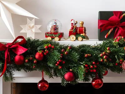 Un manteau de cheminée décoré de manière festive avec le train décoratif de la collection Christmas Toy-collection de Villeroy & Boch. Avec les ornements rouges d'une boule à neige, des décorations en forme d'étoile et une boîte cadeau verte ornée d'un ruban rouge et d'un bâton de sucre.