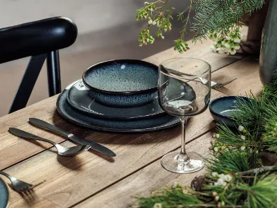 Une table en bois est dressée avec un verre Winter Glow ainsi que des assiettes et des plats creux en céramique Crafted bleu foncé. Des plantes vertes et des fleurs blanches décorent la table.
