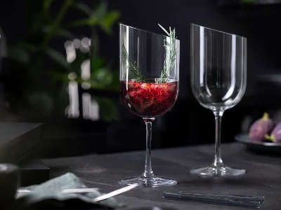 Un verre NewMoon de Villeroy & Boch contenant un liquide rouge, orné de fruits et d’une branche de romarin, posé à côté d’un verre vide sur une table sombre.