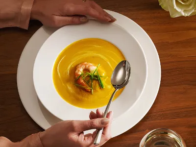 A person holds a Villeroy & Boch For Me bowl of creamy yellow soup, garnished with shrimp and herbs, on a wooden table with a glass of water and a rose.