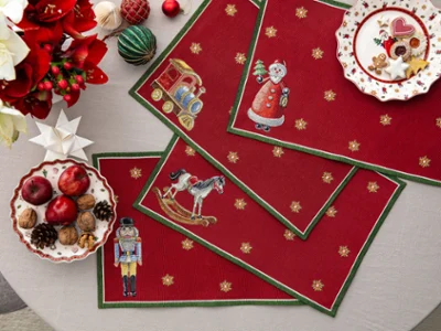 Toy's Delight Gobelin placemats with Christmas motifs Overhead view of four red Toy's Delight tapestry placemats featuring a nutcracker, rocking horse, train, and Santa Claus on a dining table, decorated with Christmas cookies, apples, walnuts, and festive ornaments
