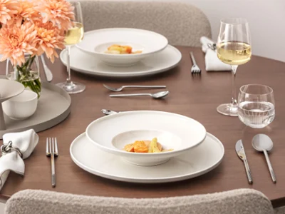 Table à manger dressée de la collection Afina Une table bien dressée de la collection Afina avec deux assiettes garnies de plats gastronomiques, des verres à vin, des verres à eau, des couverts en argent, des serviettes en tissu et un vase avec des fleurs de pêcher sur une surface en bois.