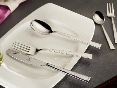 A white rectangular plate from Villeroy & Boch Blacksmith features a shiny fork, knife, and spoon on a gray surface. Next to it are a smaller spoon and a fork from the Blacksmith collection. A wine glass and a pink napkin are partially visible.