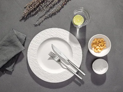 A white plate with fork and knife, a glass of water with lemon, a bowl of crackers and a napkin on a grey table.