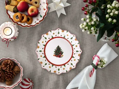 Une table dressée de manière festive avec une assiette décorée d'un motif de sapin de Noël, des biscuits, des fruits, des bougies, des serviettes et une composition florale. 
