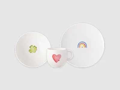 Drei weiße Teller: ein Teller mit einem Regenbogen, eine Schüssel mit einer grünen Blume und eine Tasse mit einem rosa Herz auf grauem Hintergrund.