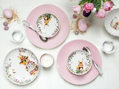 Spring Fantasy plates and decorative items on pink Rose Garden plates. Two place settings with pink plates, bowls decorated with bunny patterns, spoons, water glasses, a candle and decorative eggs from the Villeroy & Boch Spring Fantasy collection. A bunch of pink tulips can be seen on the top right.