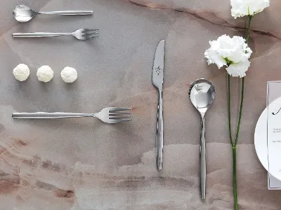 Couverts MetroChic composés d'un couteau, d'une fourchette, d'une cuillère sur une table avec trois fleurs blanches et trois petites boules blanches sur une surface en pierre à côté d'une petite assiette blanche avec des lignes noires.