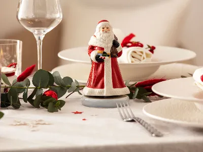 Eine festlich gedeckte Tafel mit einer Nostaligic Melody Weihnachtsmann Spieluhr in der Mitte, umgeben von Tellern, Gläsern, Besteck und Grünpflanzen.