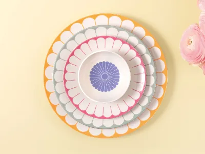 Quatre assiettes Fleur de Villeroy & Boch de différentes tailles et couleurs posées sur un fond jaune clair, à côté de fleurs roses.