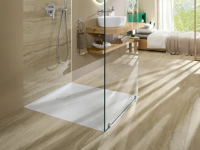 Ecora shower tray in an open walk-in shower next to a bedroom Villeroy & Boch Ecora shower tray in an open walk-in shower with travertine-look surfaces, beside a bright bedroom with a garden view.