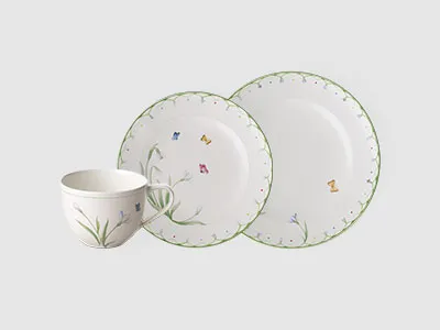 Weiße Tasse und zwei Teller mit grünem Rand und kleinen Blumenmustern auf schlichtem hellem Hintergrund.