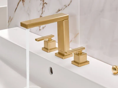 Gold Mettlach three-hole washbasin tap with running water A modern gold bathroom tap with running water, mounted on a white washbasin in front of a marble splashback.