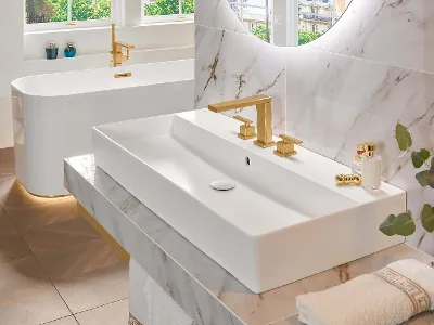 Modernes Badezimmer mit Marmoroberflächen, einem großen rechteckigen Waschbecken, goldenen Armaturen, einem runden Spiegel, einer freistehenden Badewanne und einer grünen Pflanze als Dekoration. Natürliches Licht fällt durch die Fenster.