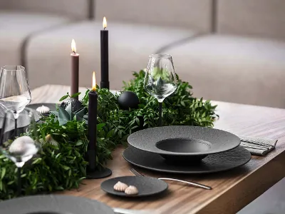Une table de salle à manger avec des assiettes noires Manufacture, des couverts de table, des verres à vin et des bougies sombres. Des plantes vertes sont utilisées comme décoration de table et créent une atmosphère raffinée.
