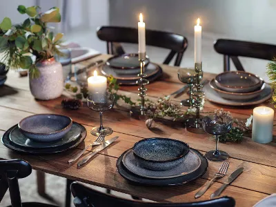 A rustic dining table with dark lava plates, burning candles in holders, glassware, flatware, and green foliage decor.