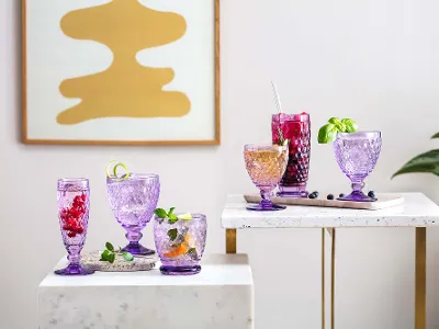 Bicchieri Boston viola di Villeroy & Boch su due tavoli bianchi con gambe dorate riempiti con delle bevande