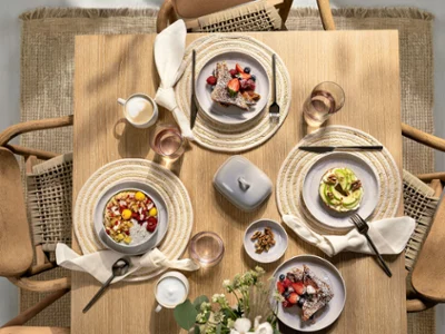 Top view of a thoughtfully arranged wooden table featuring pieces from Villeroy & Boch’s Perlemor Sand and Perlemor Home collections: plate, cereal bowl, handled mug, dip bowl, and butter dish, complemented by Like Pearl water glasses. The natural textures, soft glazes, and balanced tones create a calm, elegant dining atmosphere with a handcrafted feel.