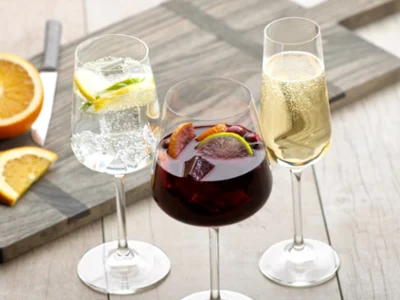 Sobre una mesa de madera hay tres copas Ovid de Villeroy & Boch: una contiene un líquido transparente con rodajas de cítricos, otra está llena de un líquido rojo oscuro y rodajas de fruta y la tercera contiene una bebida efervescente de color amarillo claro. Hay rodajas de naranja a su lado.