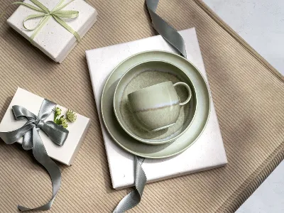 A green Perlemor cup, bowl and plate on gifts wrapped with grey ribbons, arranged on a beige textured surface.