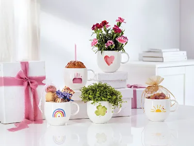 Na białej powierzchni stoi pięć białych kubków With Love marki Villeroy & Boch, każdy z innym motywem: kwiatów, słodyczy i prezentów. Czarującą scenerię uzupełniają w tle dwa prezenty owinięte różowymi wstążkami.