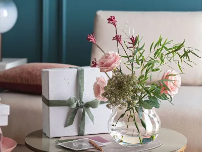 A Rose Garden vase with pink flowers and a gift on a table.