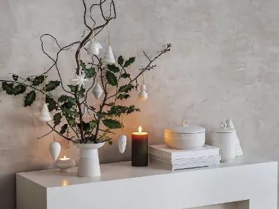 Moderne, minimalistische Weihnachtsdekoration auf einem weißen Regal mit Winter Glow Vase, Zweigen und hängenden Ornamenten. 