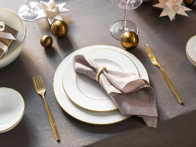 Table élégante composée d'assiettes blanches, de couverts dorés, d'une serviette beige avec un anneau, d'ornements décoratifs et de cadeaux emballés sur une nappe grise.